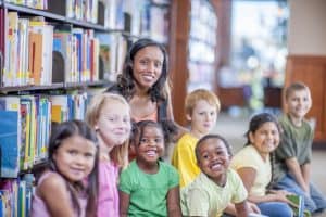 A group of elementary age students are sitting in the library with books. The children are smiling and looking at the camera.