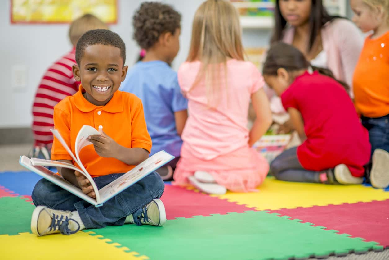 A little boy in preschool is sitting on a foam mat with his classmates and is holding a picture book - he is smiling and looking at the camera.