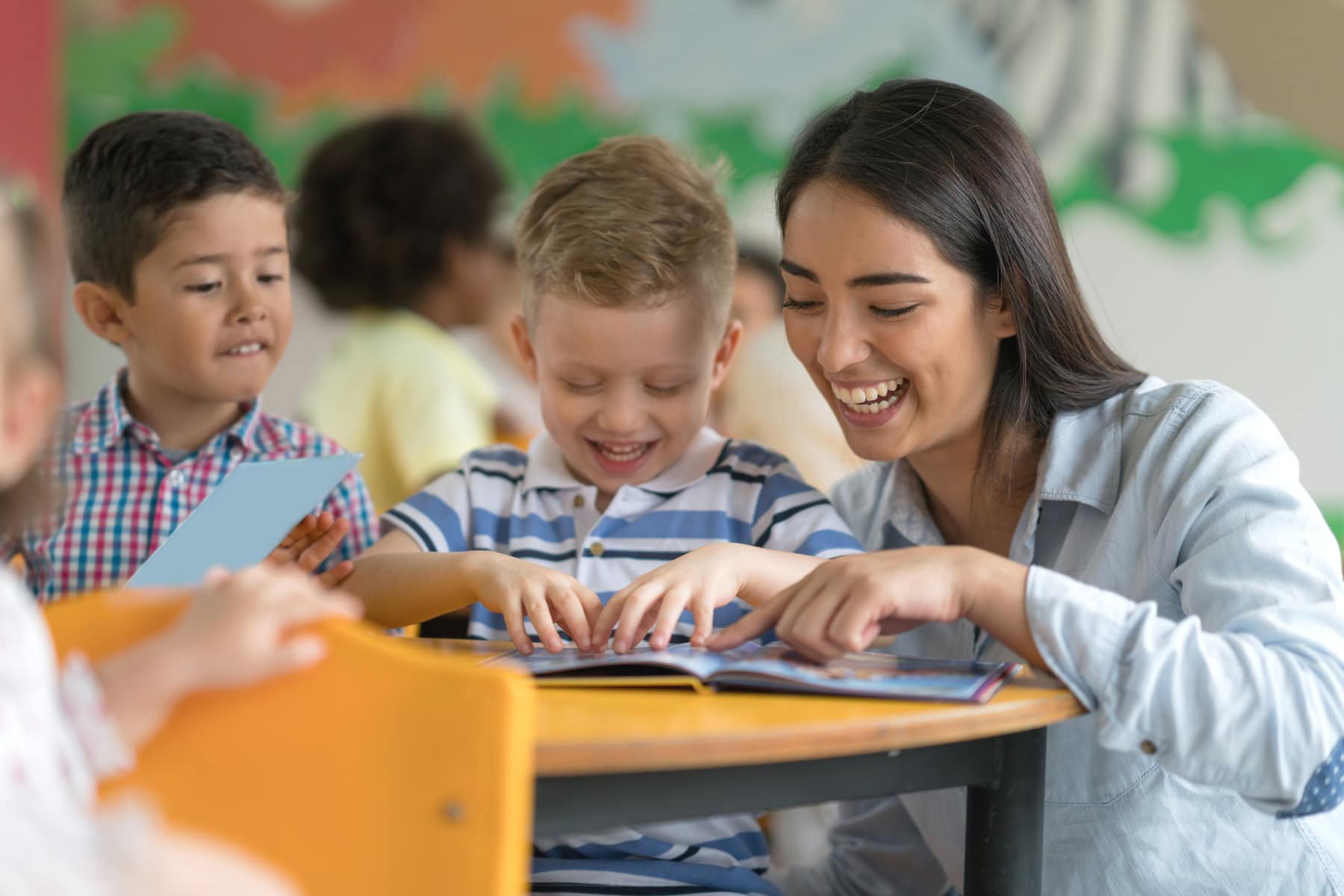 A teacher works at a table with some students. She is helping a boy read a book.