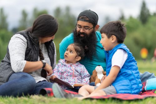 A Native American mom and dad hang out at the park and read books with their two young children on a warm summer afternoon. They're all sitting on the grass together.