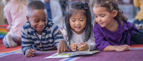 Three multi-ethnic daycare children lay close together on the floor, with a book open in front of them as they look at the pictures and read together.  They are propped up on their elbows and smiling as they enjoying their time together.  Their friends can be seen playing in the background.
