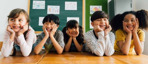 Cheerful multiethnic school friends facing camera with hands on chin, happiness, togetherness, friendship. Portrait of school children smiling in classroom